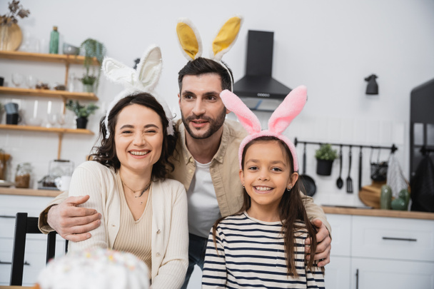 Hombre sonriente abrazando esposa e hija en diademas de Pascua en la cocina  - Foto, Imagen