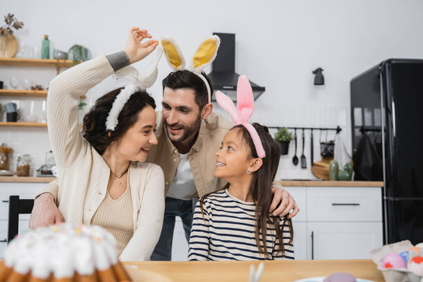 Smiling man hugging daughter and wife in Easter headbands at home  - Fotoğraf, Görsel