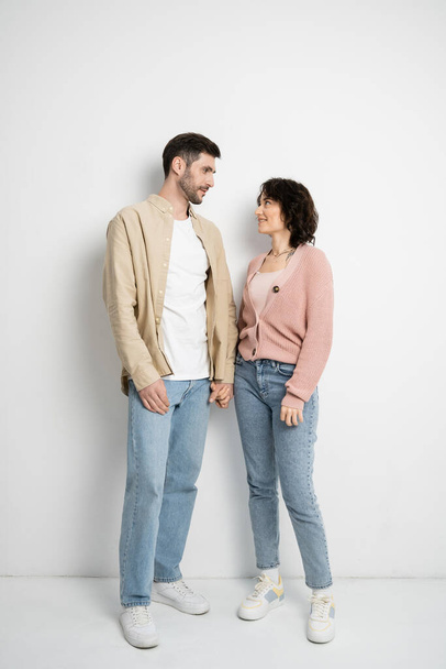 Longitud completa de la pareja sonriente cogida de la mano mientras está de pie sobre fondo blanco  - Foto, Imagen