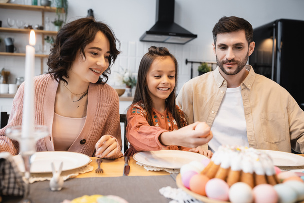 Smiling parents looking at child holding Easter egg during festive dinner at home  - Photo, Image