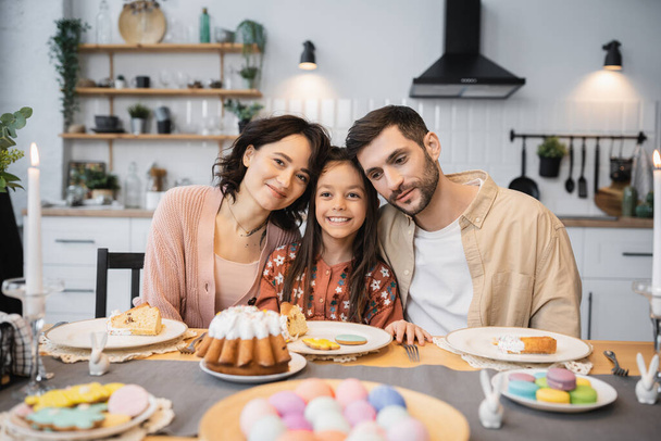 Vidám szülők és a gyerek közelében ül ünnepi húsvéti vacsora a konyhában  - Fotó, kép