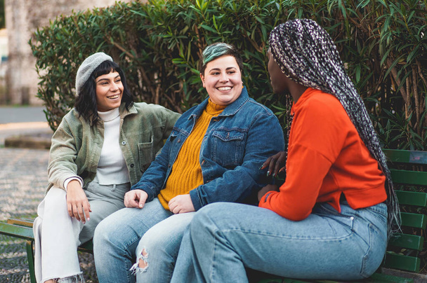 Три женщины, две кавказки (одна брюнетка, одна пышная и небинарная) и одна африканка с дредами в волосах, сидят вместе на скамейке в парке, беседуют - Фото, изображение