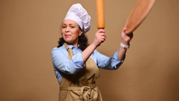 Joyful female pastry chef in professional uniform, moving to music, singing and dancing with wooden cutting board and rolling pin, over beige background. Baking. Culinary master class concept - Felvétel, videó