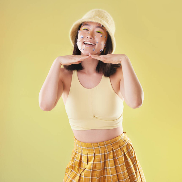 Fashion, hands on face and portrait of woman with comic eyes isolated on yellow background. Happy, funny and asian girl with a smile for gen z beauty and creativity with color clothes in studio. - Fotoğraf, Görsel