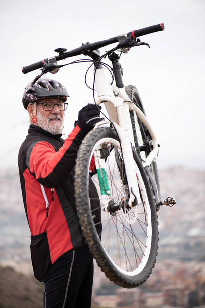 Porträt eines älteren Rentners in Radlerausrüstung, der mit dem Fahrrad in der Luft fährt, die Stadt zu seinen Füßen. - Foto, Bild