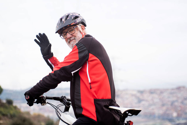 retired elderly male cyclist with bike and equipment waving, city in the background - Fotoğraf, Görsel