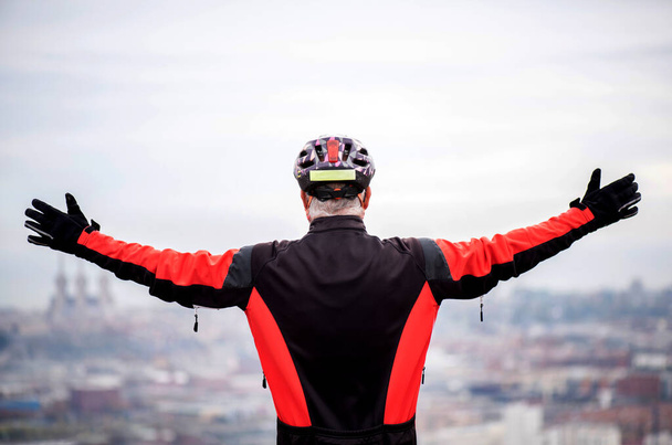 older man dressed as a cyclist with helmet on his back with his arms in the air with symbol of freedom and life, at his feet the city of barcelona - Photo, Image