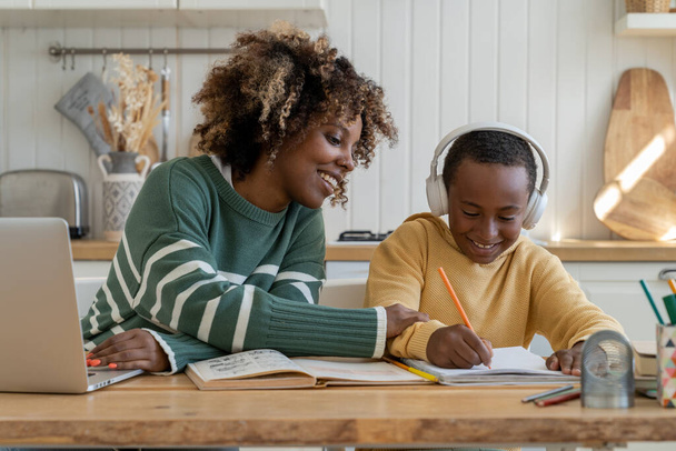 Joyful African American mom praises pleased son in headphones for success in studies and creative art task, distracted from remote work as freelance with laptop sitting together at home kitchen table - Photo, image