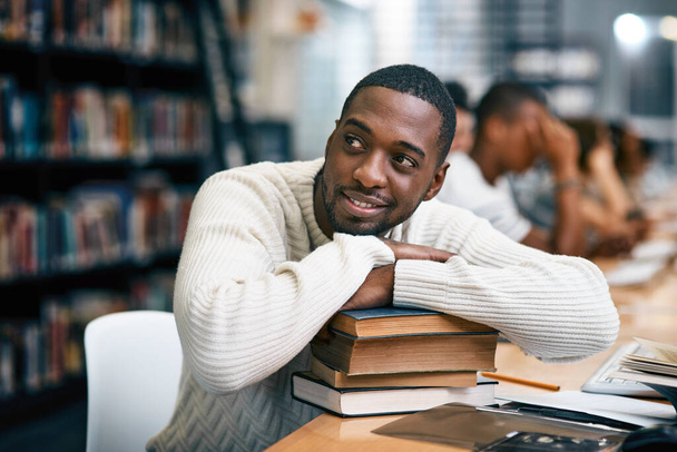 Mentale noot om het zelf te laten gebeuren. een jonge man rust op een stapel boeken in een universiteitsbibliotheek en kijkt bedachtzaam - Foto, afbeelding
