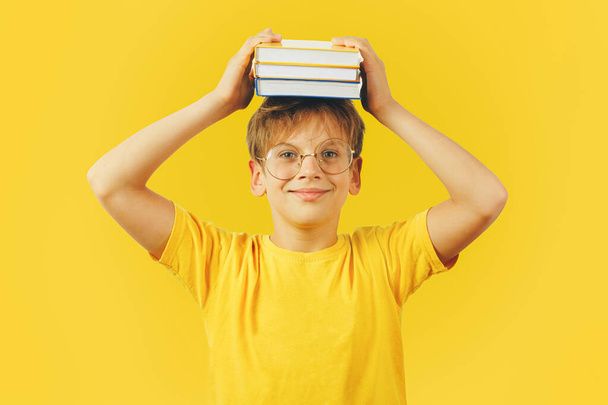 Il ragazzo in età scolare con gli occhiali tiene libri di testo in testa e sorride. Vestita con T-shirt gialla senza iscrizioni. Studio girato su sfondo giallo. Concetto di istruzione. - Foto, immagini