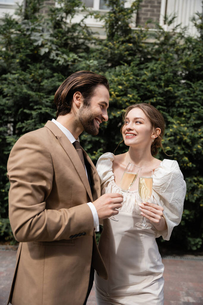 smiling groom in suit and happy bride in wedding dress clinking glasses of champagne  - Fotografie, Obrázek