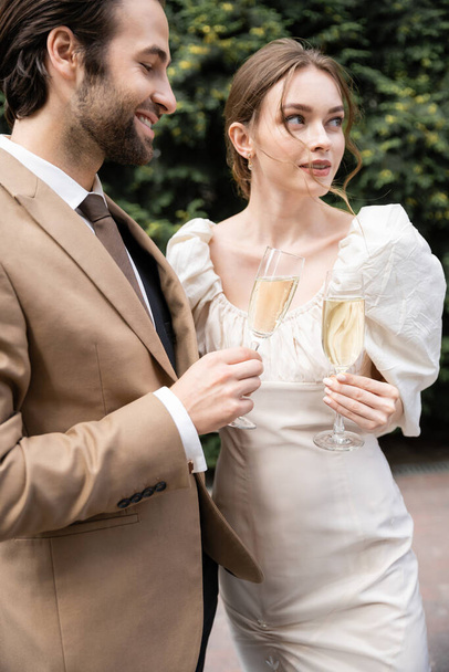 glücklicher Bräutigam im Anzug hält Glas Champagner mit junger Braut im Brautkleid  - Foto, Bild