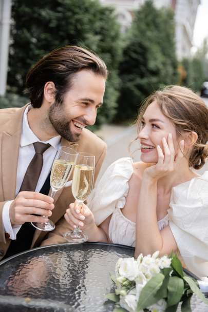 веселые и молодые молодожены звонят в бокалы шампанского, улыбаясь во время свадебного торжества  - Фото, изображение