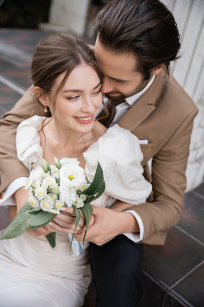 vue aérienne du marié barbu embrassant mariée souriante en robe blanche avec bouquet de mariage  - Photo, image