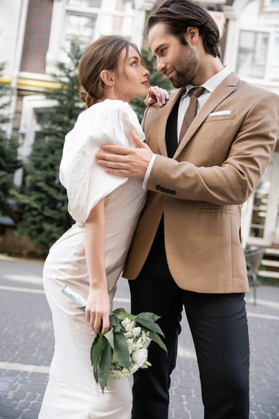 bearded groom in suit hugging young bride in white dress with wedding bouquet while standing on street  - Foto, afbeelding
