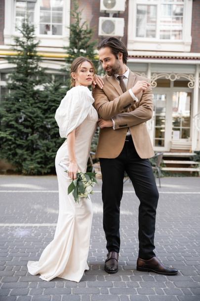 full length of young bride in white dress holding wedding bouquet while standing near cheerful groom in suit  - Fotografie, Obrázek