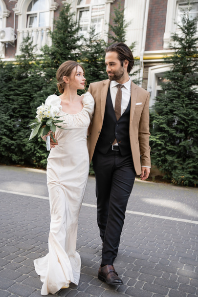 full length of gorgeous bride in white dress holding wedding bouquet while standing with bearded groom on street  - Photo, image