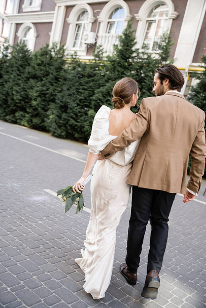 back view of bride in white dress holding wedding bouquet and walking with groom on street  - Foto, immagini