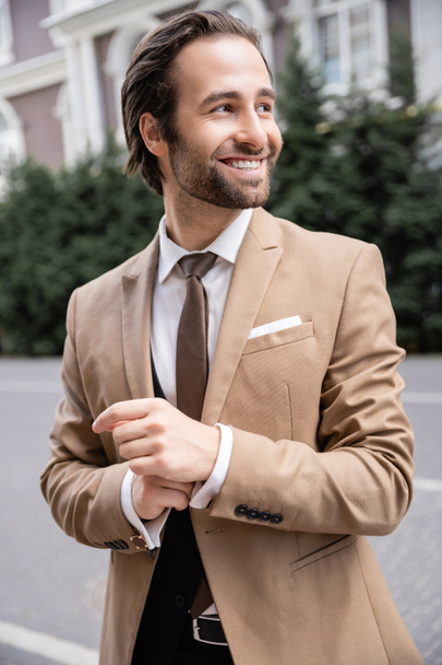 cheerful groom in beige formal wear adjusting sleeve on shirt  - Photo, image