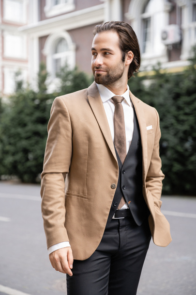 young groom in beige formal wear standing with hand in pocket and looking away on street  - Fotografie, Obrázek