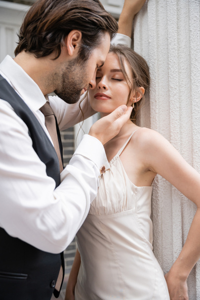 bearded groom in formal wear touching face of pretty bride with closed eyes  - Zdjęcie, obraz