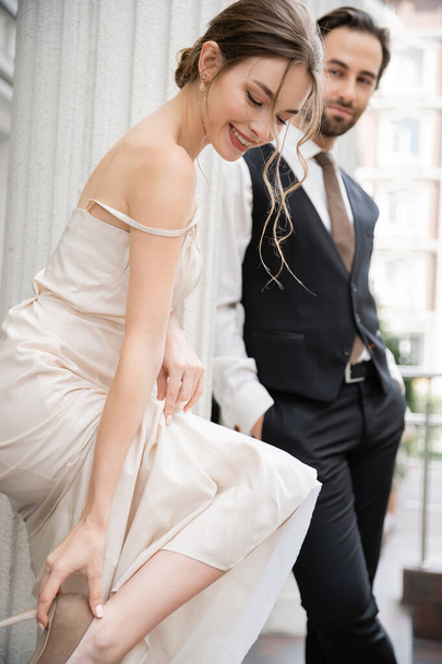 счастливая невеста в свадебном платье в туфлях на высоком каблуке рядом с женихом в формальной одежде на размытом фоне  - Фото, изображение
