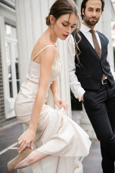 pretty bride in wedding dress wearing high heeled shoe near groom on blurred background  - Foto, Imagem