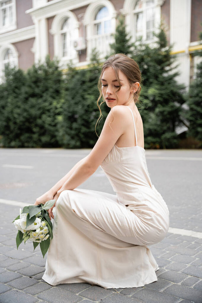 young bride in wedding dress holding bouquet with flowers while sitting outside  - Photo, Image