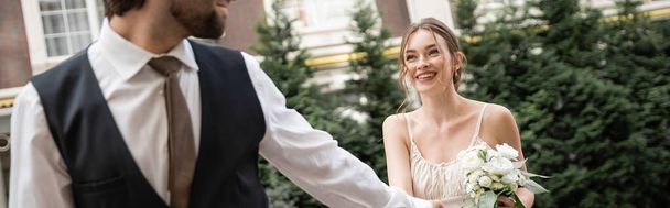 happy bride in white dress with wedding bouquet holding hands with groom while walking outside, banner  - Fotografie, Obrázek