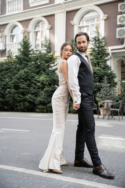 full length of young bride and groom in wedding dress and suit holding hands outside  - Fotó, kép