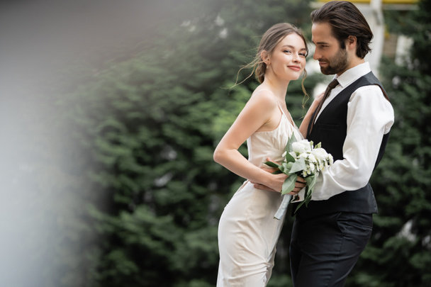 bearded groom in vest embracing happy bride in white dress with wedding bouquet  - 写真・画像
