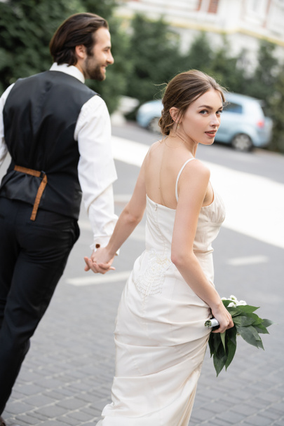 groom holding hand of gorgeous bride in wedding dress with bouquet of flowers  - Photo, Image