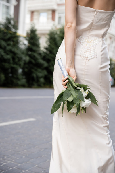 cropped view of young woman in white dress holding wedding bouquet behind back  - 写真・画像