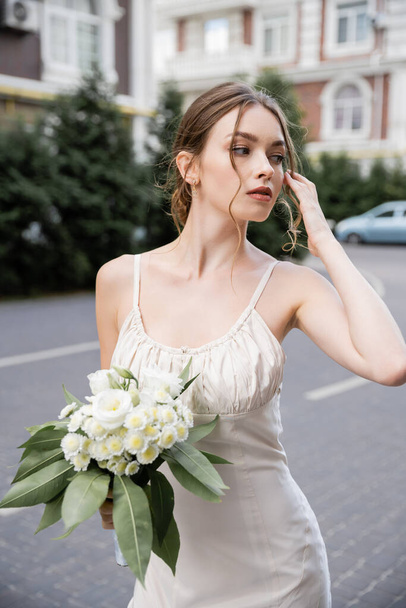 young woman in wedding dress holding bouquet with flowers and looking away - Foto, afbeelding