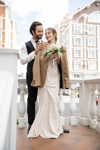 full length of happy groom in vest hugging cheerful bride in beige blazer and wedding dress with bouquet  - Foto, imagen