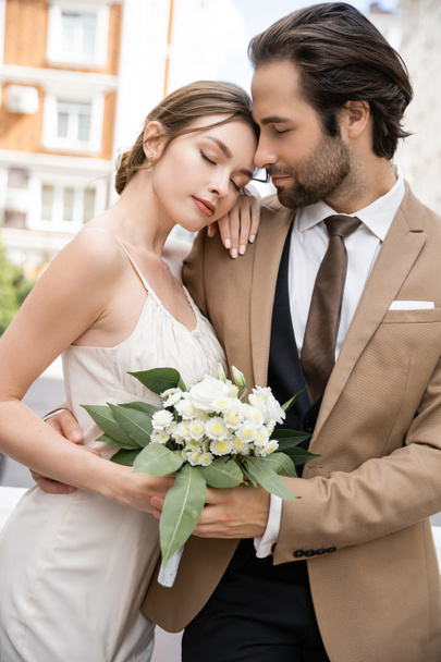 young bride in wedding dress holding bouquet and leaning on groom in suit  - Photo, image