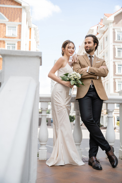 full length of happy young bride in wedding dress holding bouquet and leaning on groom in suit  - Fotó, kép