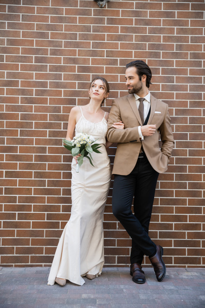 full length of dreamy bride in wedding dress holding flowers near groom in suit while standing against brick wall  - Fotoğraf, Görsel