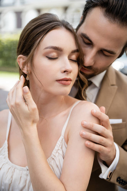 man met baard in pak met stropdas knuffelen mooie bruid met gesloten ogen buiten - Foto, afbeelding