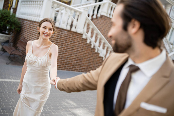 blurred groom in suit holding hand of happy bride in white dress  - Foto, Bild
