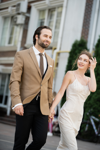 happy bride in wedding dress looking at groom while holding hands on street  - 写真・画像