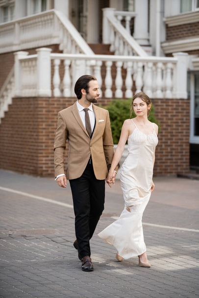 повна довжина нареченої у весільній сукні тримає руки з нареченим, тримаючи руки на вулиці
  - Фото, зображення