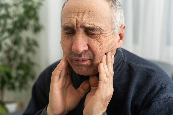 Old man with toothache. Elderly senior man has toothache. Unhappy man face in tooth pain sitting on sofa at home, feel sick unwell. Sad aged man hand holding his chin. Adult suffering toothache. - Foto, immagini