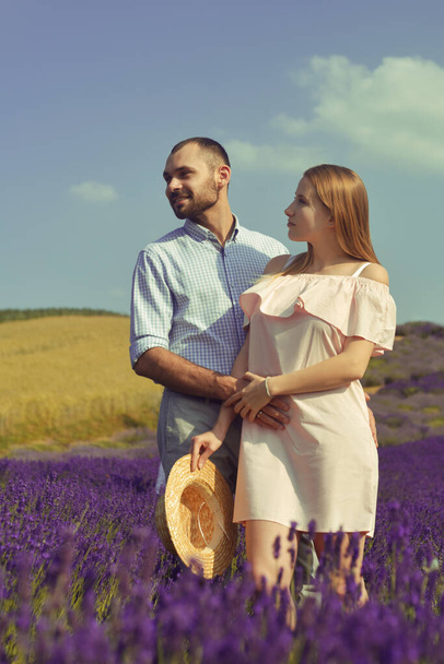A couple in love in a field of lavender at sunset in good weather. Beautiful woman in a dress and straw panama with a man on the background of nature, love and feelings. Blue, purple lavender - Zdjęcie, obraz