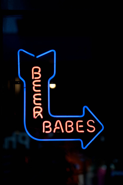Close-up on a blue neon light shaped into an arrow pointing right with a reon "Beer babes" inside. - Fotoğraf, Görsel