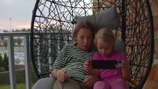 Αδέλφια συγκόλληση πάνω από το παιχνίδι σε φορητή κονσόλα σε μια κουνιστή καρέκλα στο μπαλκόνι. 11-year-old αγόρι και 5-year-old αδελφή του κάθονται μαζί σε μια ζεστή καρέκλα swing στο μπαλκόνι τους και απορροφάται σε μια διασκέδαση - Πλάνα, βίντεο