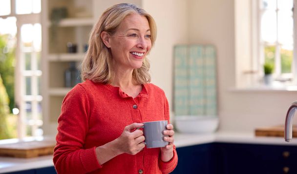 Smiling Mature Woman Standing In Kitchen Relaxing With Cup Of Coffee - Φωτογραφία, εικόνα