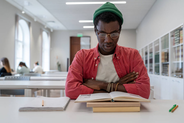 Könyv fókuszált afro-amerikai diák srác olvassa tanulmányi információkat keresztbe tett karokkal felkészülve a nehéz vizsgára az egyetemi könyvtárban. Gondos ember szemüvegben olvasott tankönyv nehéz feladat - Fotó, kép