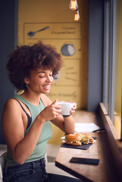 Female Customer In Coffee Shop Window Eating Bagel Sandwich For Lunch - Foto, immagini