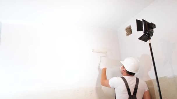 Молодий чоловік в шоломі і однорідний малює білі стіни в квартирі валиком, використовуючи лампу і прожектор для освітлення. Сучасний ремонт в квартирі
 - Кадри, відео